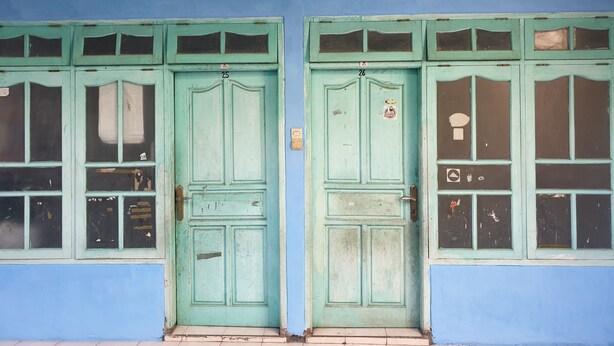 Fachada de edificio con dos puertas verdes, numeradas 25 y 26, y varias ventanas que muestran signos de desgaste.