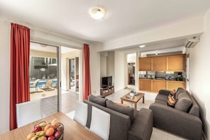 A modern living room with a sofa set, a dining table with fruit, and a kitchen in the background. - Villa Coral Bay Sun (Pegeia)