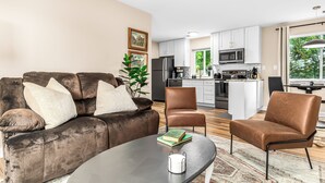 A modern living room with a brown leather sofa, two brown armchairs, and a coffee table. A kitchen with white cabinets and a dining area with a table and chairs is visible in the background.