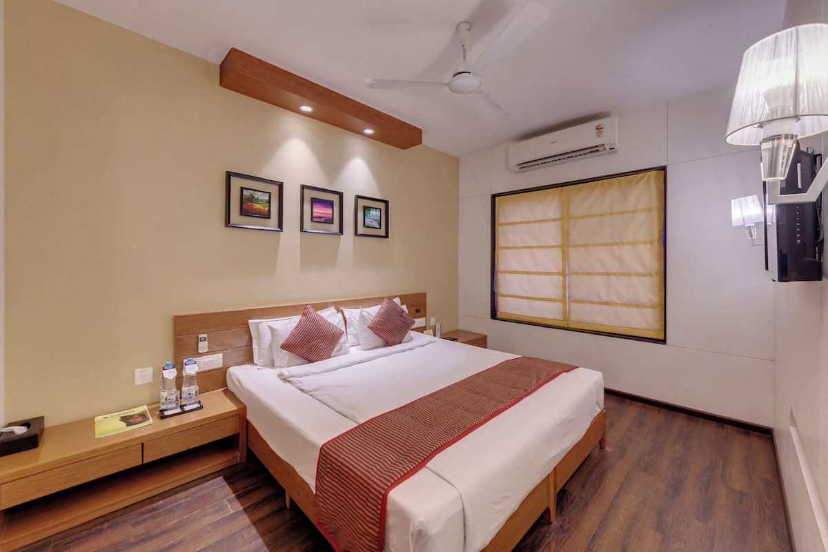 A hotel room with a bed, wooden headboard, bedside tables, framed pictures on the wall, an air conditioning unit, and a window with a curtain.
