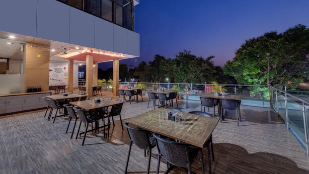 An outdoor dining area with tables and chairs, a wooden deck, and a modern building in the background. - Grand Fortune Beacon Ichalkaranji (Hatkalangda)