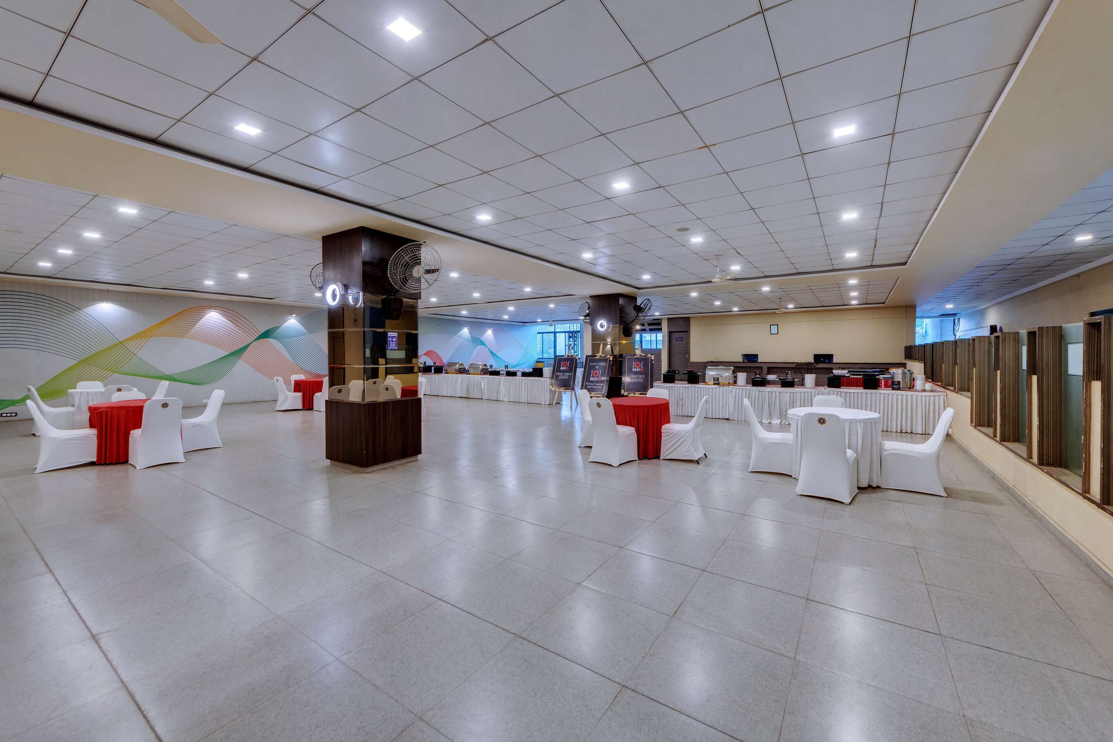 Un espacio interior amplio con varias mesas redondas y sillas blancas, una zona de recepción central y un mural grande en la pared.