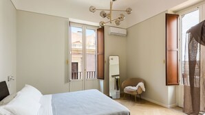 A bedroom with a bed, a chair, an air conditioning unit, and a window with a view of buildings.