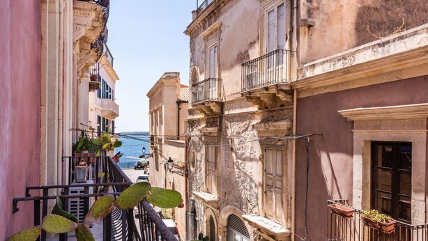 A narrow street with buildings featuring balconies and windows, overlooking a body of water.