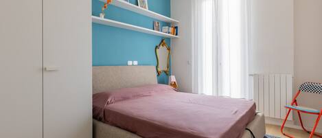 A bedroom with a bed, a chair, shelves with books, and a mirror.