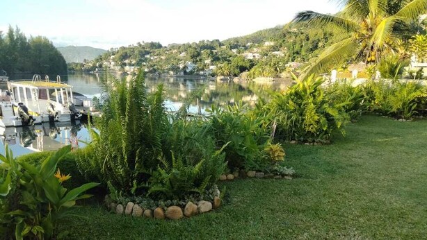 A serene lakeside garden with a wooden bench, lush greenery, and a boat docked by the water. - Mae Water Front Apartments (Mahé Island)