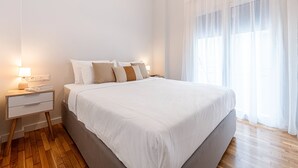 Un lit bien fait, avec une literie blanche et beige, une table de chevet avec une lampe, et une grande fenêtre ornée de rideaux légers.