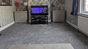 Een woonkamer met een flatscreen televisie op een standaard, grijs tapijt, behang met patroon en een raam met gordijnen.