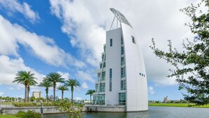 A modern building with a unique curved roof, surrounded by a water body and palm trees. - Unbelievable View- No other unit has a view like this one. (Indialantic)