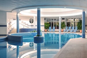 A large indoor swimming pool with a curved design, surrounded by lounge chairs and potted plants.