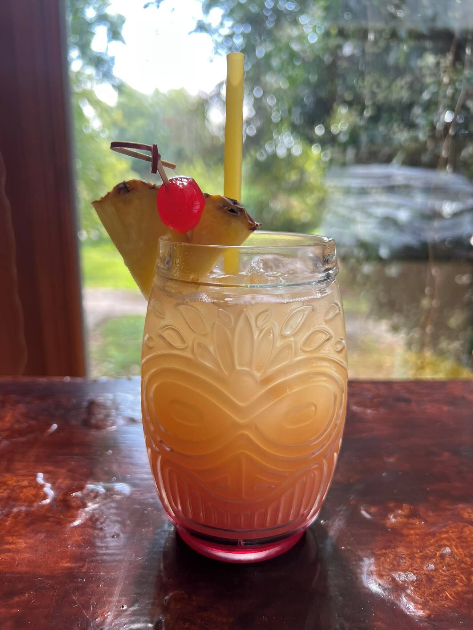 A glass with a yellow straw and a red cherry on a pineapple slice, placed on a wooden surface.