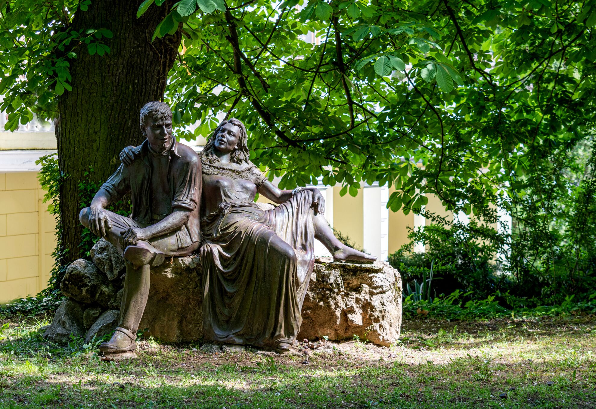 A statue of two figures seated on a rock under a tree.