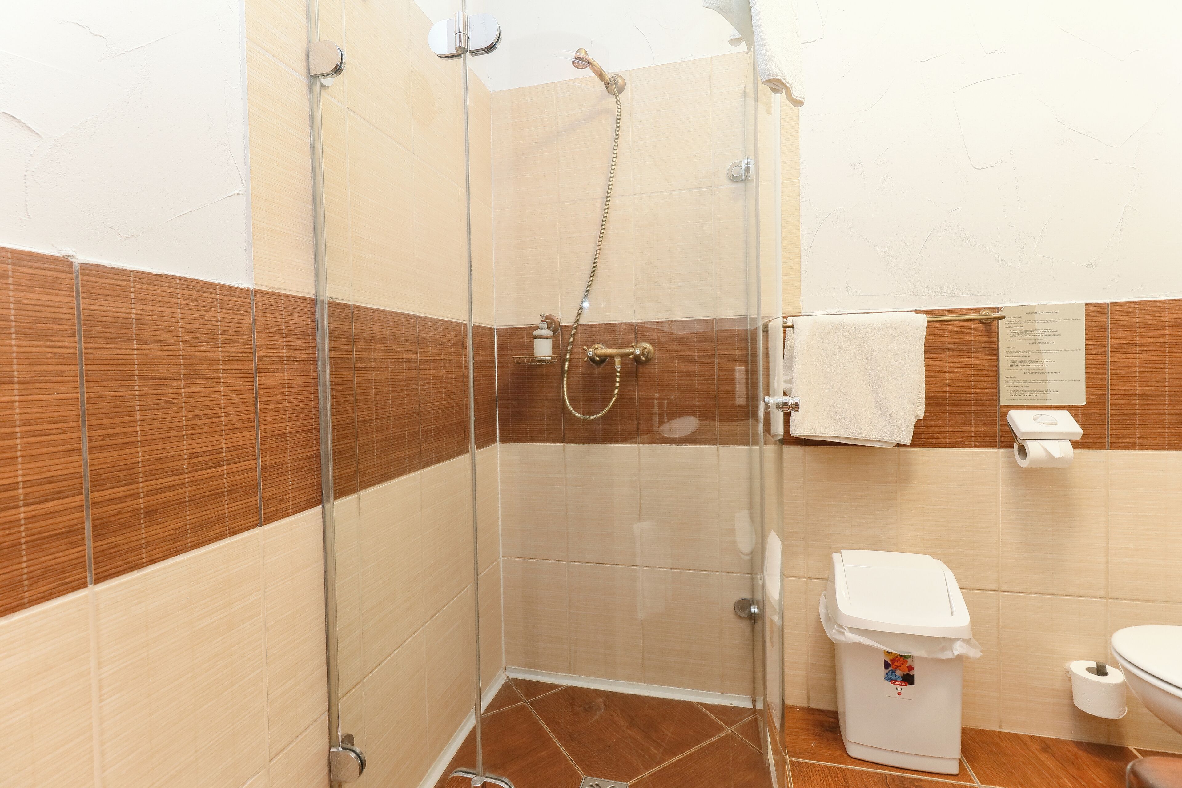Ducha con mampara de vidrio, cabezal de ducha de mano, inodoro y un cubo de basura en un baño con paredes de azulejos.
