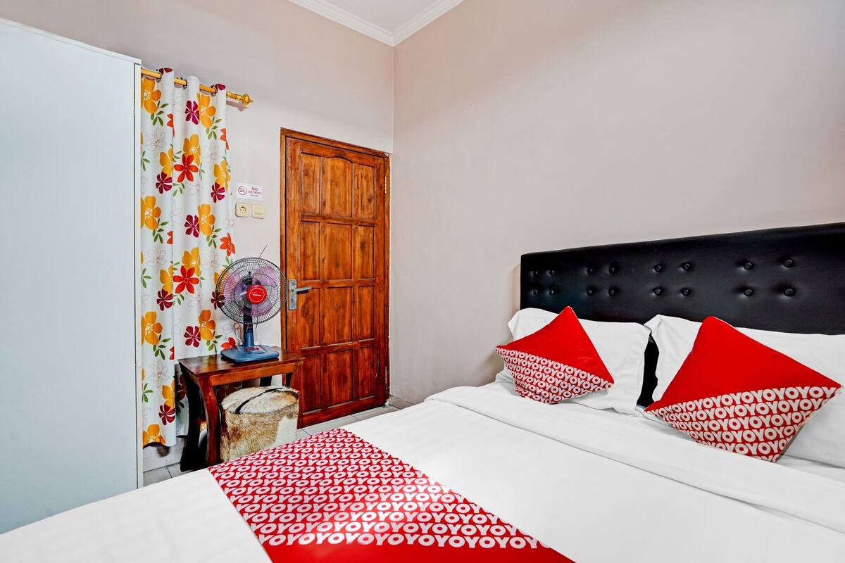 Un dormitorio con cama, armario de madera, ventilador y cortina estampada con motivos florales.