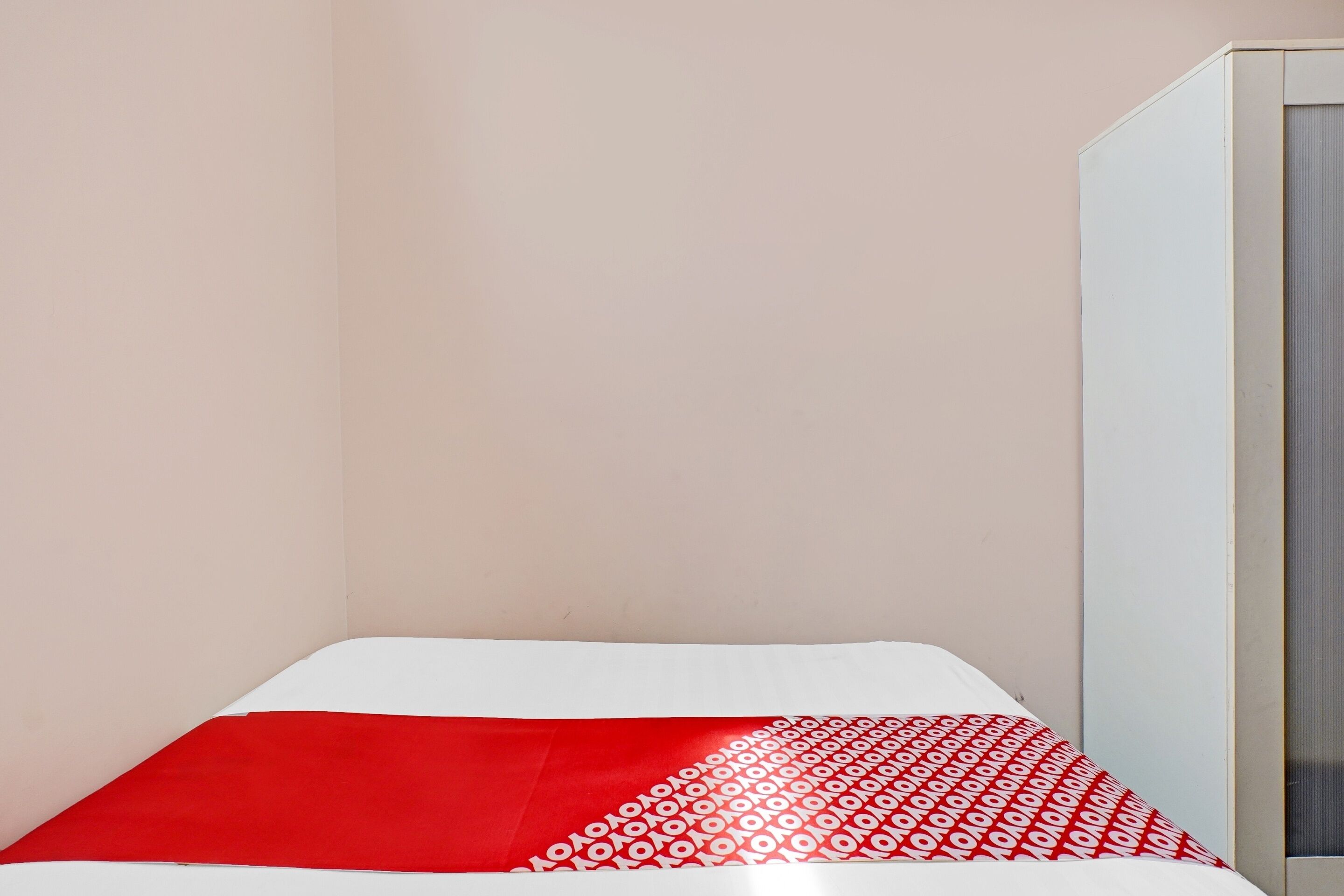 Uma cama de solteiro com colcha estampada em vermelho e branco, cabeceira branca e guarda-roupa branco em um quarto com paredes lisas.