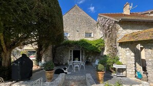 A stone building with a tiled roof, a paved courtyard with a table and chairs, and potted plants.