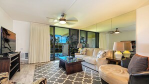 A modern living room with a sofa, coffee table, and a TV. There is a ceiling fan, a large window, and a view of the outdoors. - Kamaole Sands 10-205  Ocean View, Fountain View, Great Location, Great Rates! (Kihei)