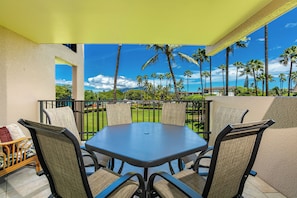 A covered outdoor seating area with a table and chairs, overlooking a landscaped garden with palm trees and a clear blue sky.