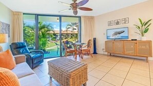 A hotel room with a large sliding glass door leading to a balcony with a view of palm trees and the ocean. There is a ceiling fan, a television, and a wicker ottoman.