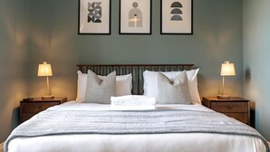 Ein ordentlich bezogenes Bett mit einer gemusterten Decke, zwei Nachttischlampen und drei gerahmten Kunstwerken an der Wand.