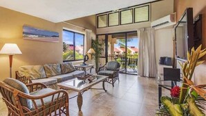A spacious living area with a sofa set, chairs, and a coffee table. There is a large window with a view of palm trees and a building. - Kamaole Sands #2-401 Beautifully Renovated Suite, Family Friendly, A/C! (Kihei)