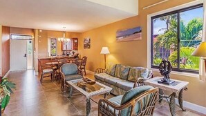 A spacious living area with a dining table, chairs, a sofa, and a coffee table. There is a large window with a view of greenery outside. - Kamaole Sands #2-401 Beautifully Renovated Suite, Family Friendly, A/C! (Kihei)