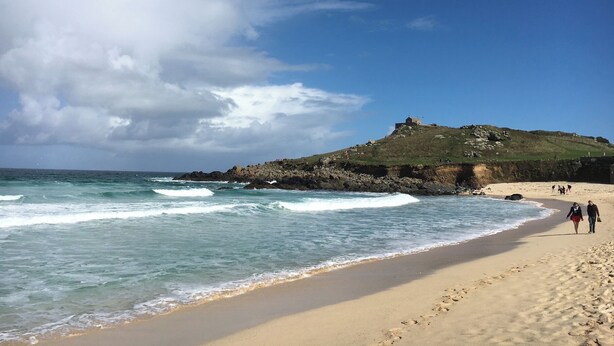 Uma praia com ondas, uma colina com uma construção e pessoas caminhando.
