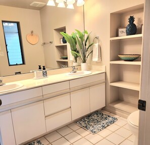 Un bany modern amb un mirall gran, mobles blancs, un lavabo, un vàter i una petita estant amb objectes decoratius.
