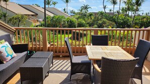 Eine überdachte Terrasse im Freien mit Korbmöbeln, einem Holztisch und Blick auf einen üppigen Garten mit Palmen.