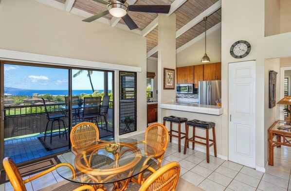 A spacious dining area with a glass table and wicker chairs, a kitchen with modern appliances, and a large sliding glass door leading to a balcony with a view of the ocean. - Wailea Ekolu #306 Panoramic Ocean Views on Wailea Golf Course, Wifi, A/C (Wailea)