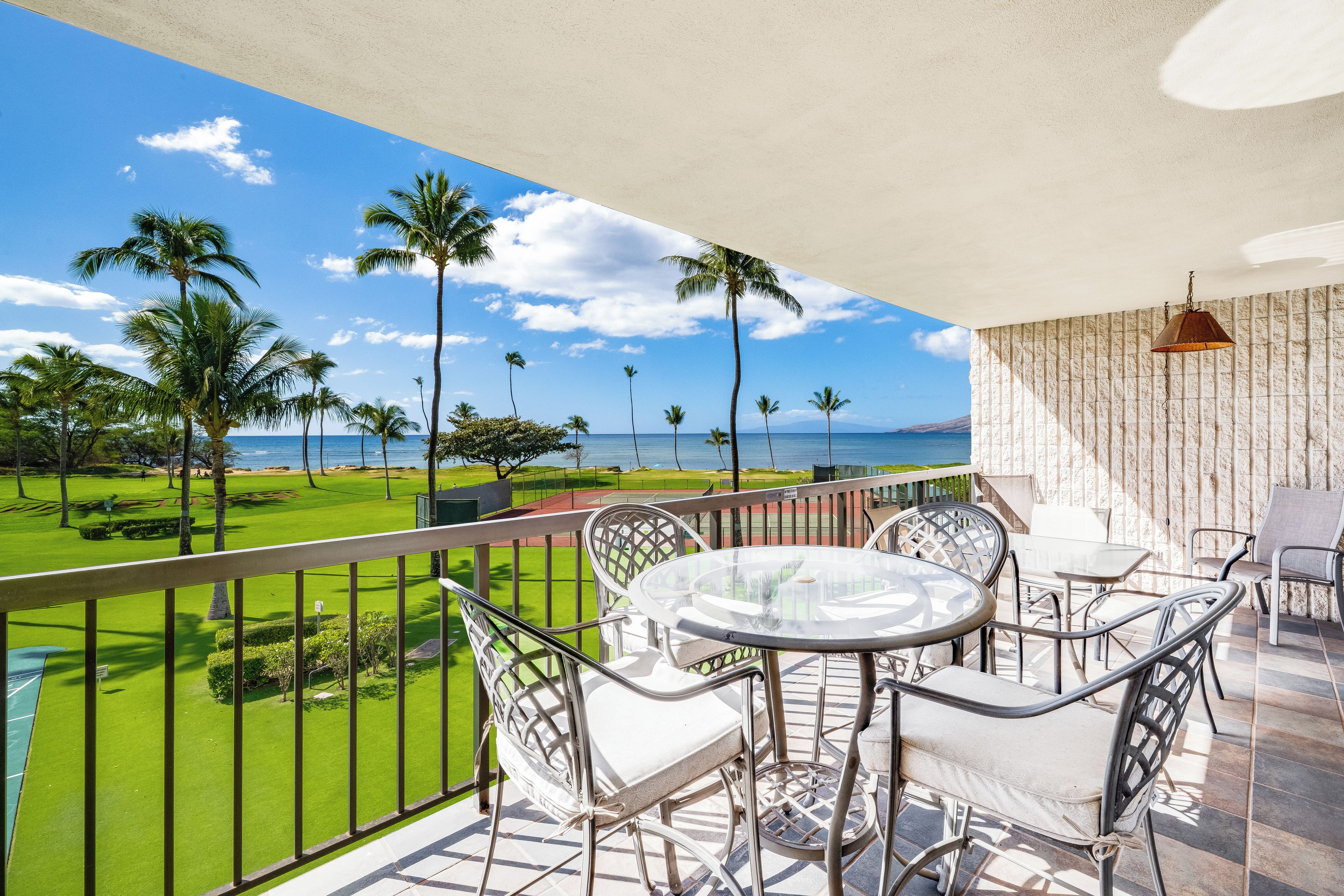 A balcony with a view of the ocean and palm trees, furnished with metal chairs and a glass table.