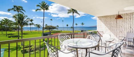 A balcony with a view of the ocean and palm trees, furnished with metal chairs and a glass table.