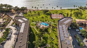 Vista aérea de un resort con piscina central, rodeado de céspedes cuidados y palmeras, con una playa y el océano al fondo.
