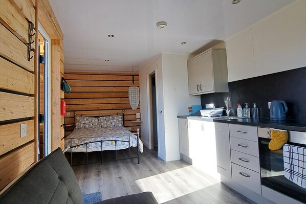 A compact bedroom with a bed, a small seating area, and a kitchenette with cabinets and a sink. - The Posh Bothy Spean Bridge (Roybridge)