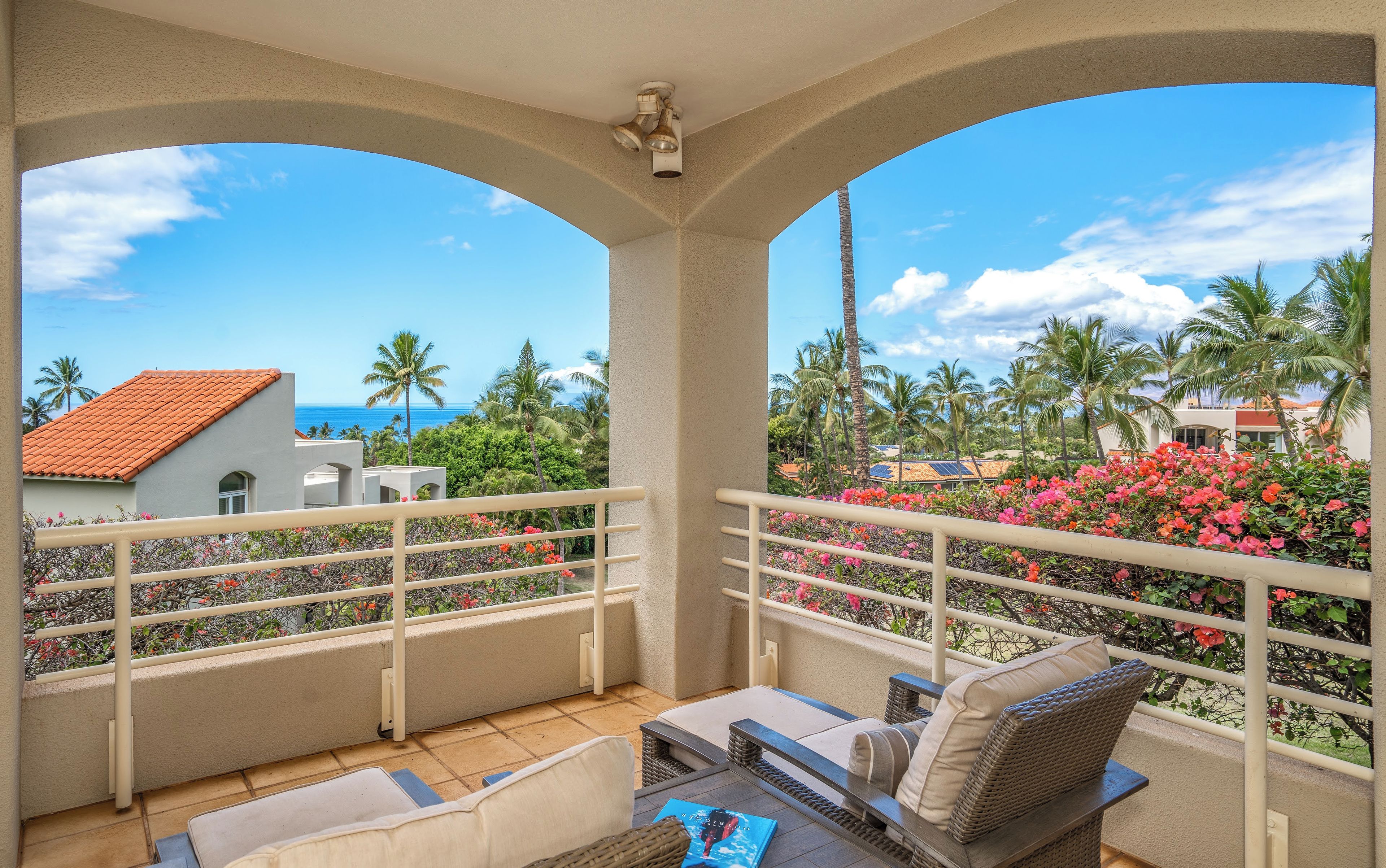 A balcony with a view of the ocean, palm trees, and a building with a red roof.