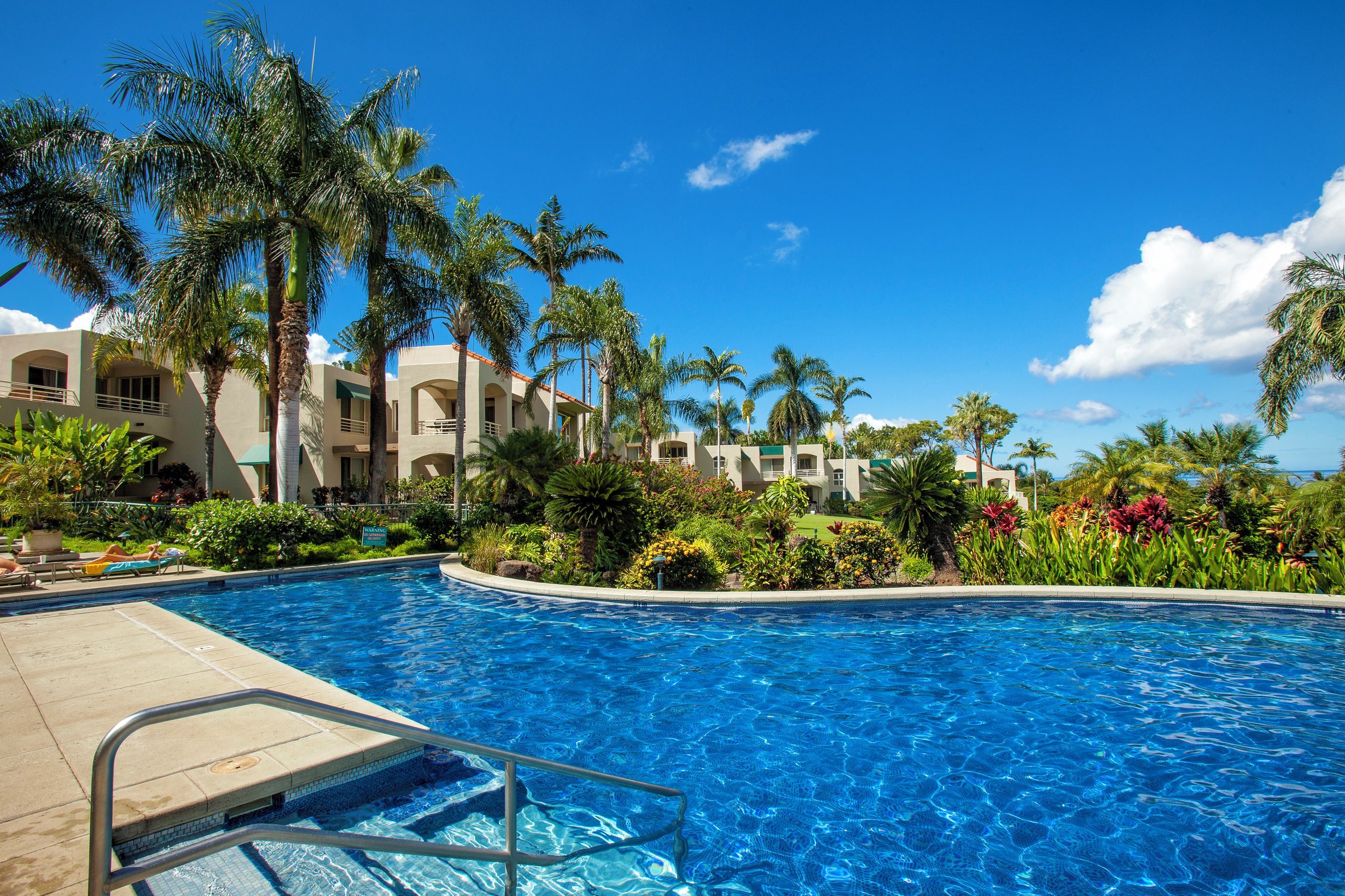 A swimming pool with a clear blue water, surrounded by palm trees and a building with multiple balconies.