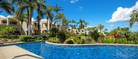 A swimming pool with a clear blue water, surrounded by palm trees and a building with multiple balconies.