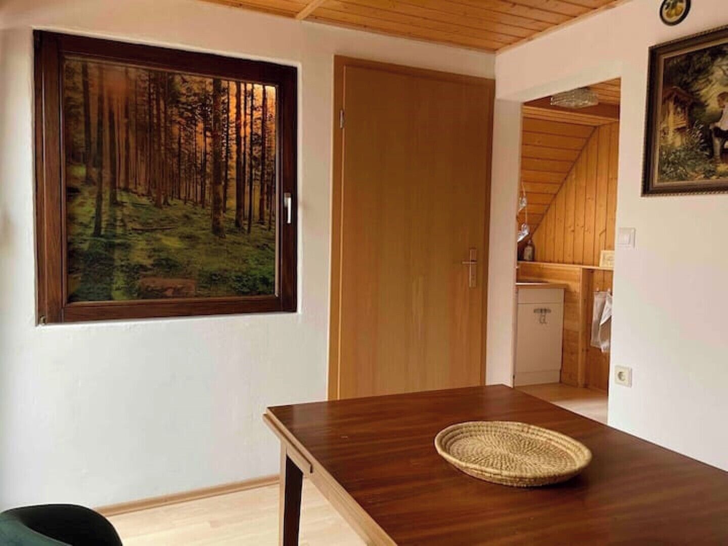 A room with a wooden table, a painting of a forest, and a door leading to another room.