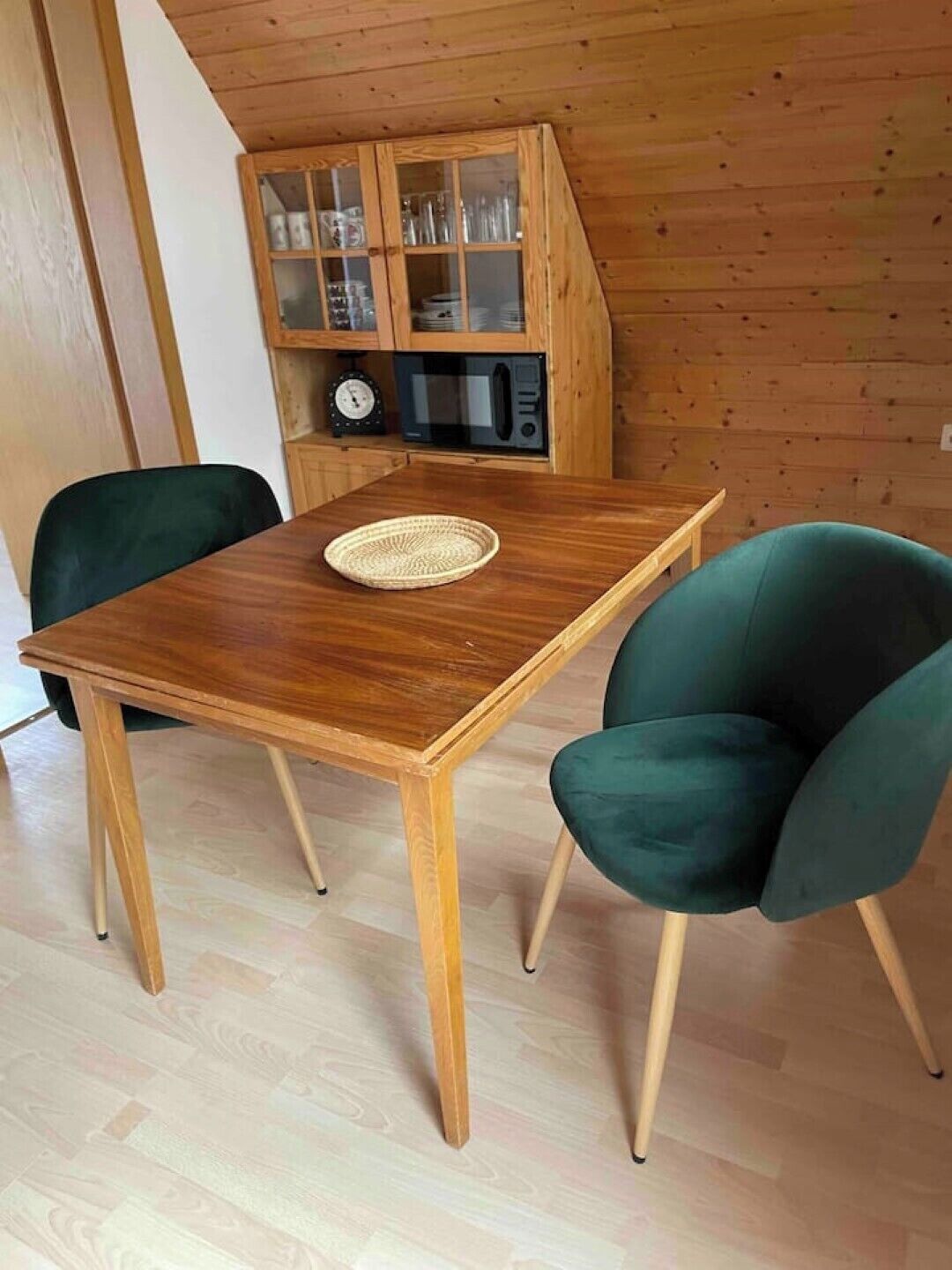 Una mesa de comedor de madera con dos sillas verdes, un mantel tejido y un microondas encima.