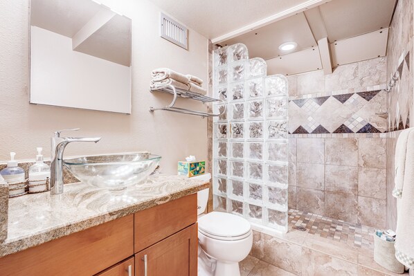 Une salle de bain avec un lavabo en verre, des toilettes, une douche avec un mur carrelé et un miroir au-dessus du lavabo.