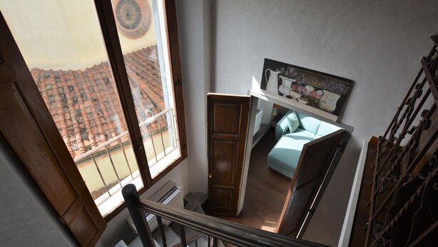 A room with a view of a tiled roof outside, a wooden door, a bed with a blue cover, and a wall-mounted clock. - Elegant Apartment for 3 in Firenze With Elevator (Florence)