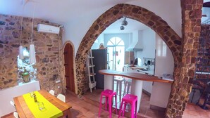 Un coin repas avec une table en bois, un chemin de table jaune et des tabourets roses. On aperçoit un mur en pierre et un espace cuisine à l’arrière-plan.