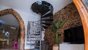 Un escalier en colimaçon avec une rampe en métal, un mur en pierre, un téléviseur posé sur un meuble blanc et une plante en pot.