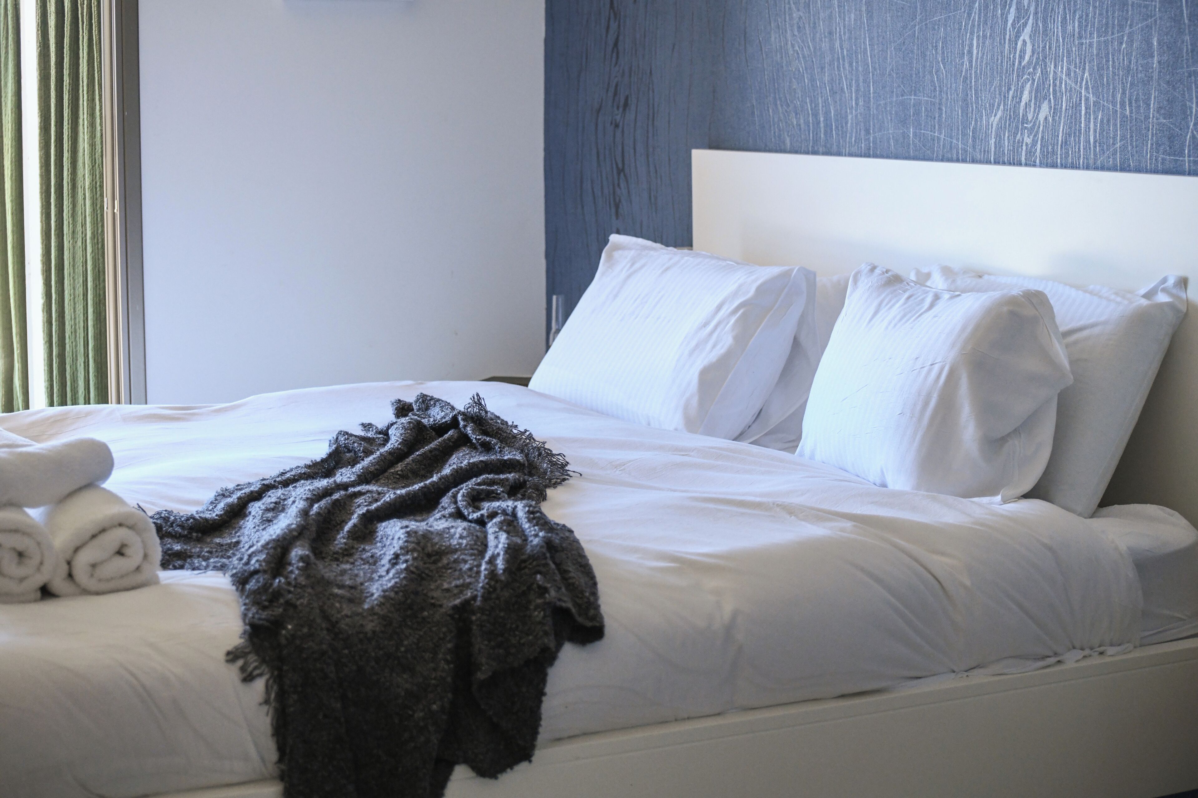 Una cama bien hecha con sábanas blancas y una manta gris oscuro, un juego de toallas enrolladas y un cabecero frente a una pared azul.