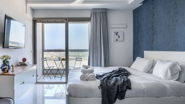 Habitación de hotel moderna con una cama grande, vistas a la playa, una zona de comedor y un televisor en la pared.