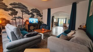 Ein Wohnzimmer mit einer Couch, einem Sessel und einem Couchtisch mit einer Kerze. Ein Fernseher steht auf einem Schrank und an der Wand befindet sich ein Wandbild mit Bäumen.