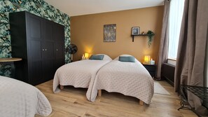 Une chambre avec deux lits, une armoire, un ventilateur, une étagère avec une plante et une fenêtre avec des rideaux.
