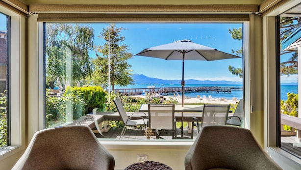 A balcony with two chairs, a woven ottoman, and a table with an umbrella. A scenic view of a pier and mountains is visible through the glass.