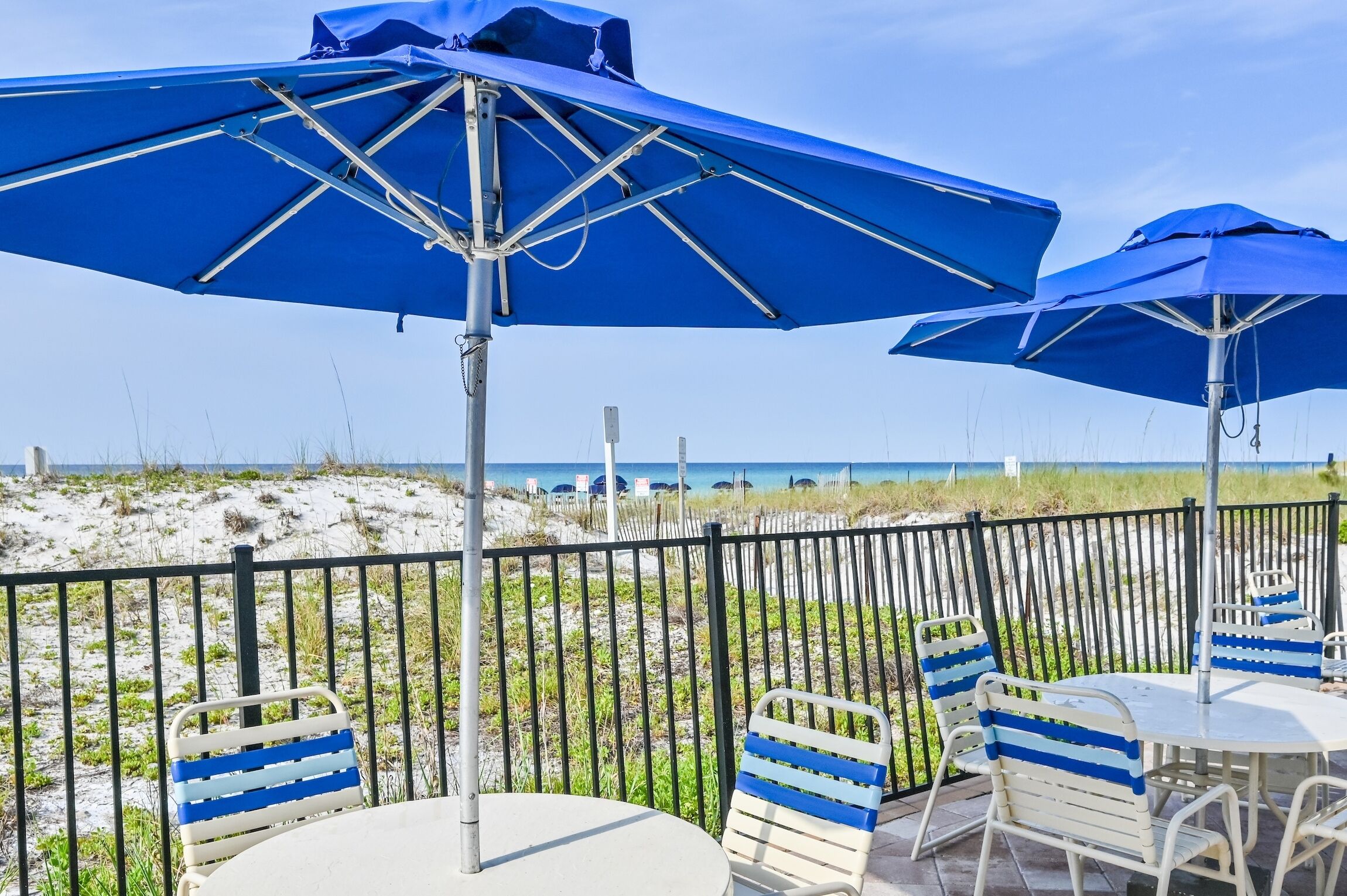 A beachside patio with blue umbrellas, white tables, and striped chairs.