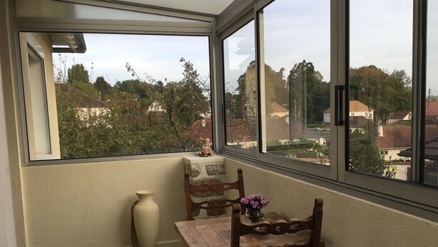 Ein Eckzimmer mit großem Fenster, einem kleinen Tisch mit Stühlen, einer Vase und einem dekorativen Gegenstand auf der Fensterbank.